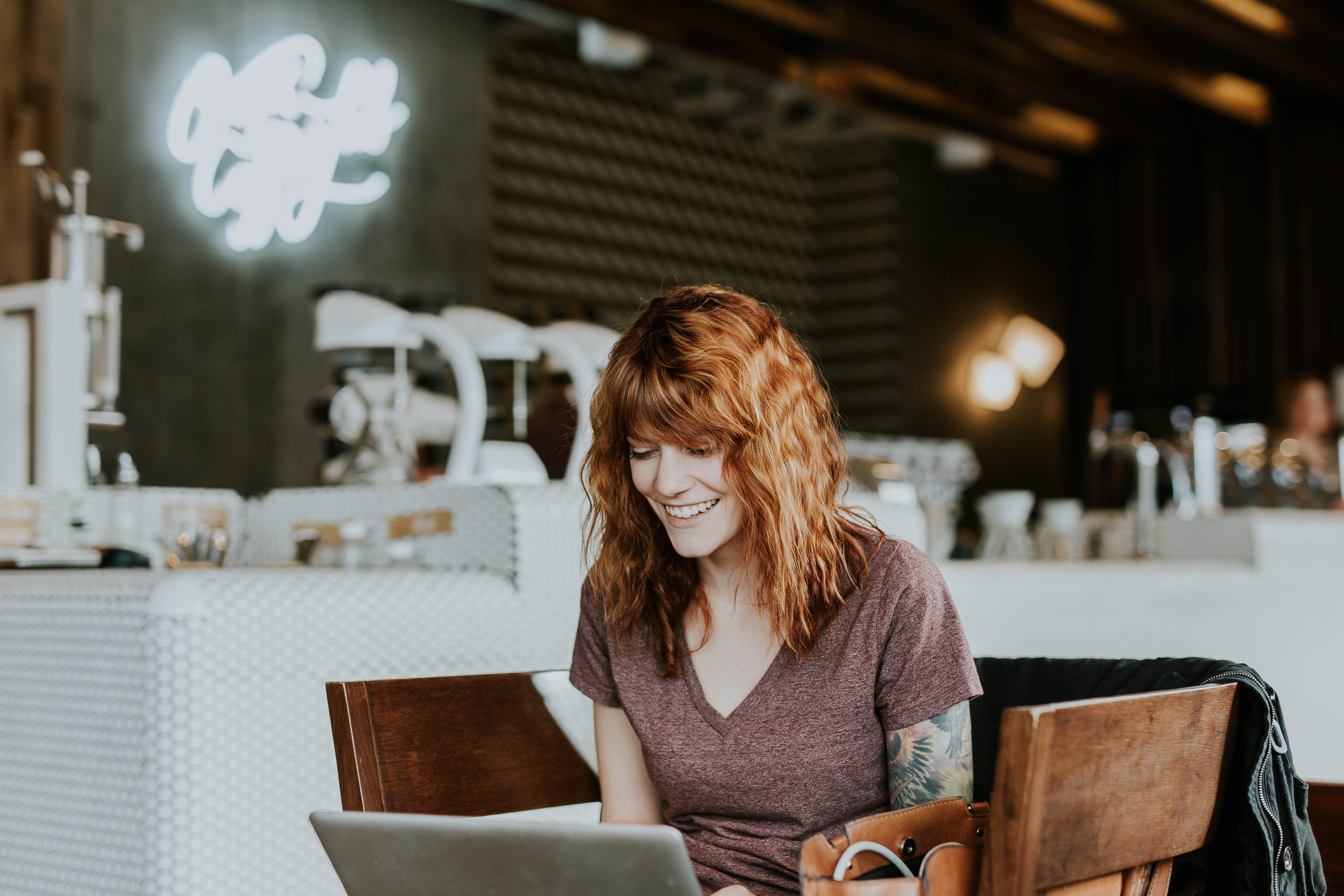Smiling red haired woman working on silver laptop with brown chairs